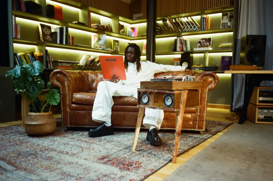 Dans un salon, un homme assis sur un canapé en cuir tient la pochette du vinyle, une platine au premier plan
